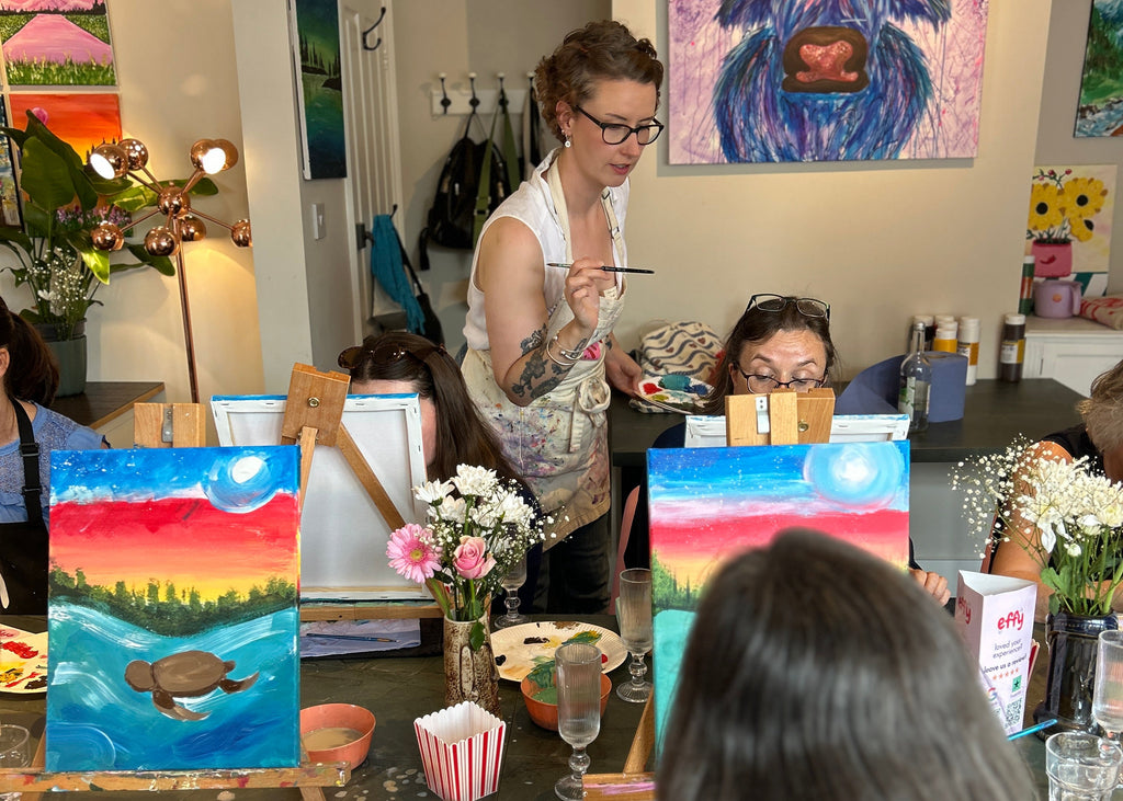A group of people participating in an indoor painting workshop or class, with several canvases featuring colorful sunset landscapes and a large blue-and-purple cow painting on display. A woman stands at the back holding a paintbrush, while others sit at tables with paint palettes, brushes, and flowers.