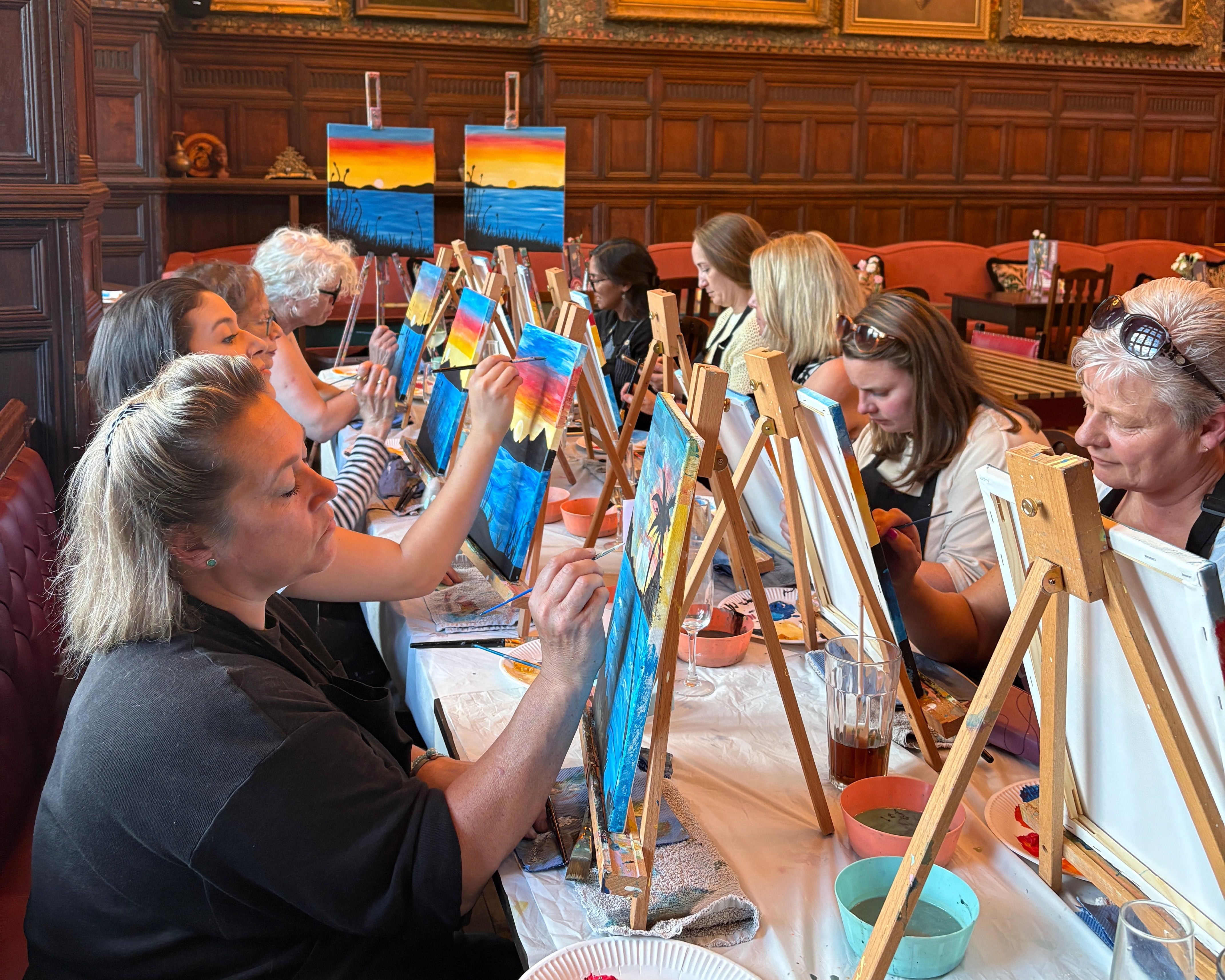 A group of people painting in a large, ornate room with wood-paneled walls and framed paintings. Several easels and paint palettes are visible on tables covered with white cloths.