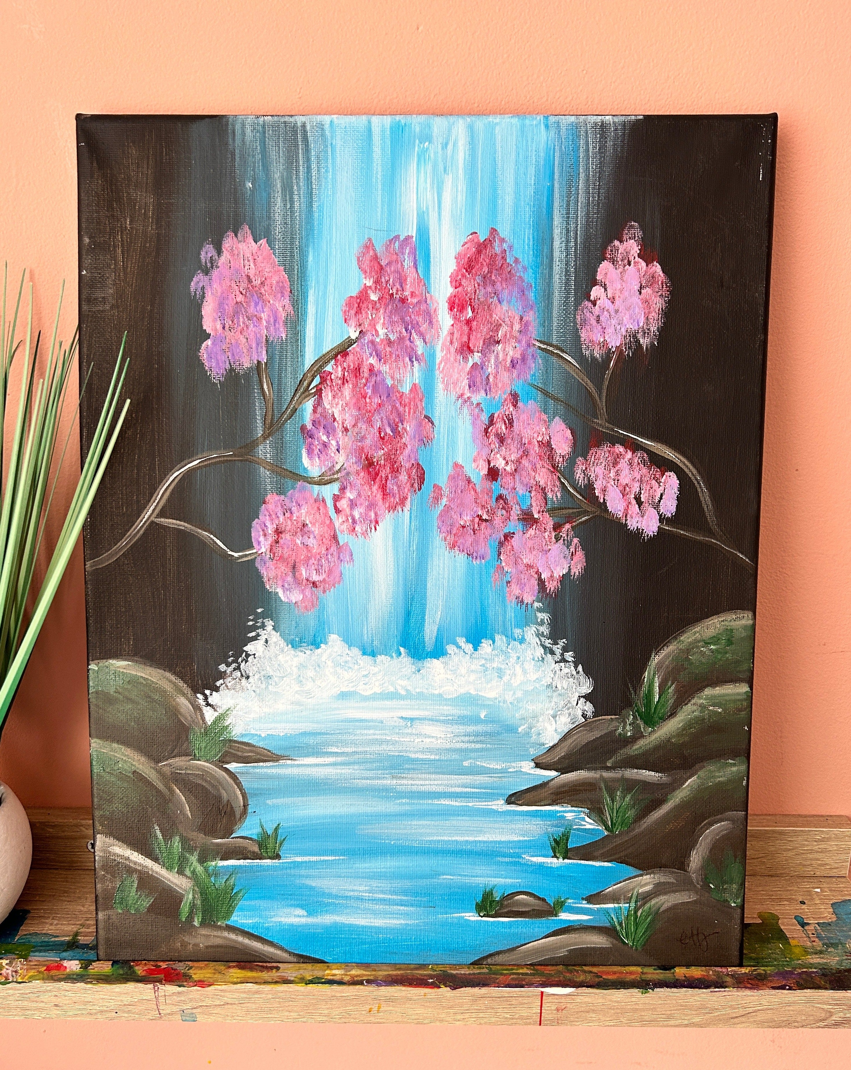 Acrylic painting of a waterfall, with pink cherry blossoms in front of it. The water heads towards the viewer of the art with grassy rocks surrounding the edge of the canvas. 