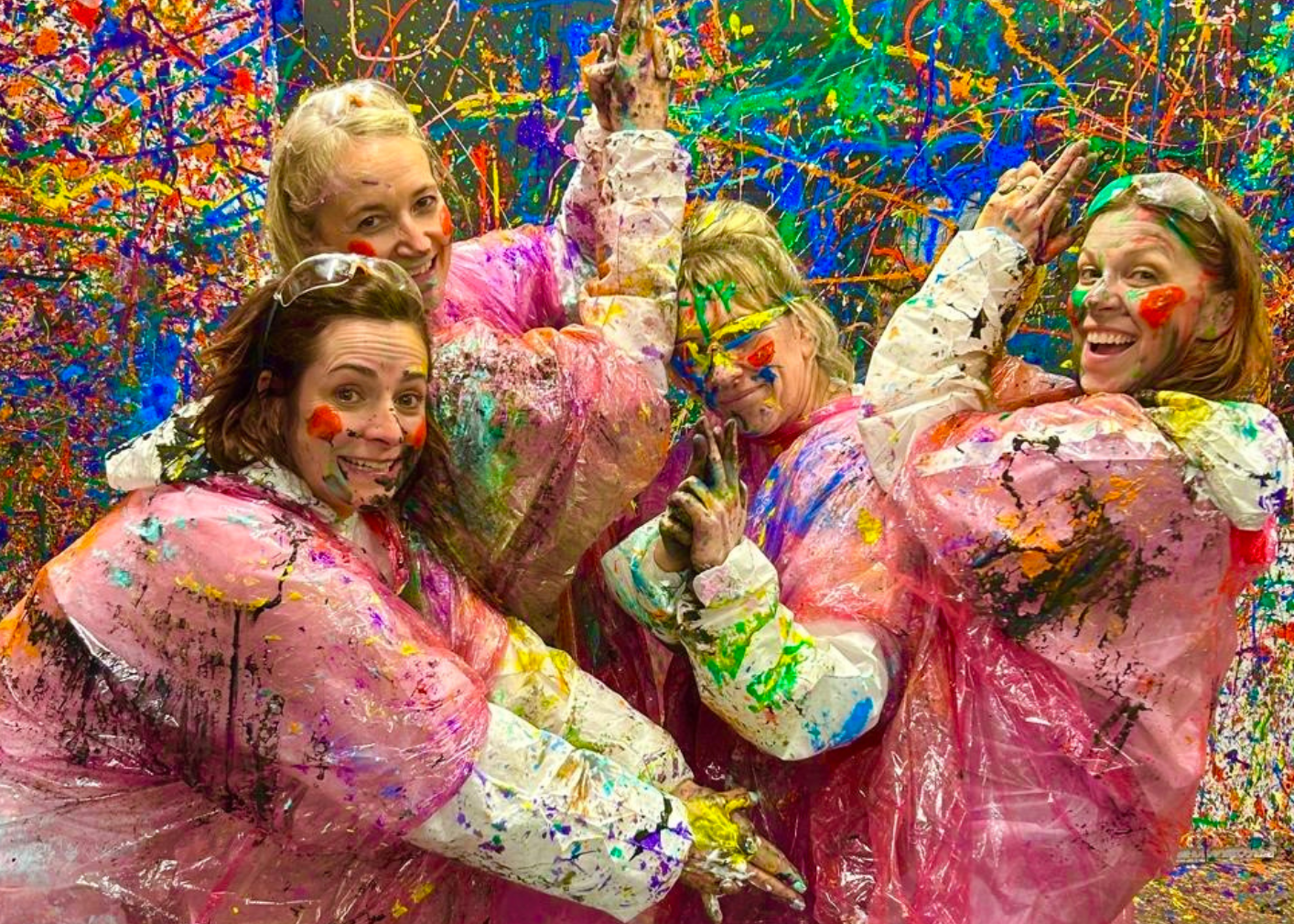 Four women wearing pink plastic paint splatter suits and white pants pose together in a paint-splattered room, with colorful paint drips and splatters covering the background.