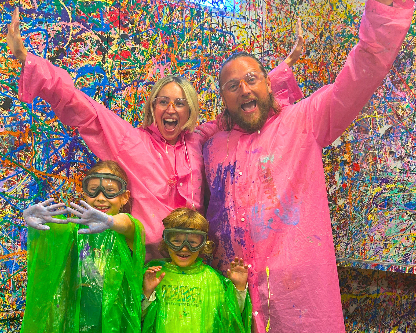 A family of four posing inside a colorful spray-paint-covered room, wearing bright pink and green plastic poncho rain suits and protective face masks.